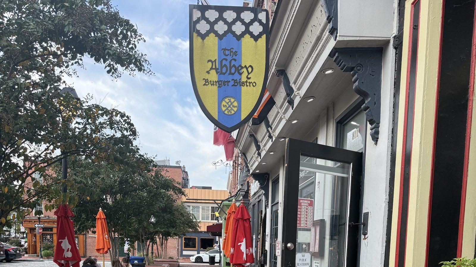 The Abbey Burger Bistro's shield sign over its door on Broadway in Fells Point, with picnic tables and the square behind
