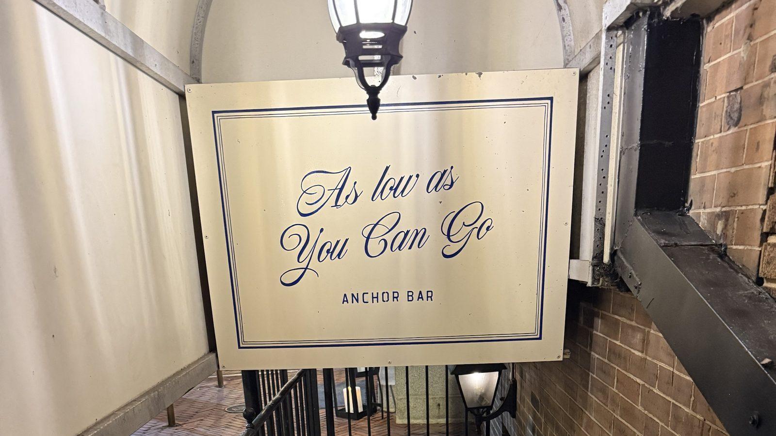 The Anchor Bar's hanging sign, reading 'As low as You Can Go', lit by a lamp over the railed stairwell down to the bar