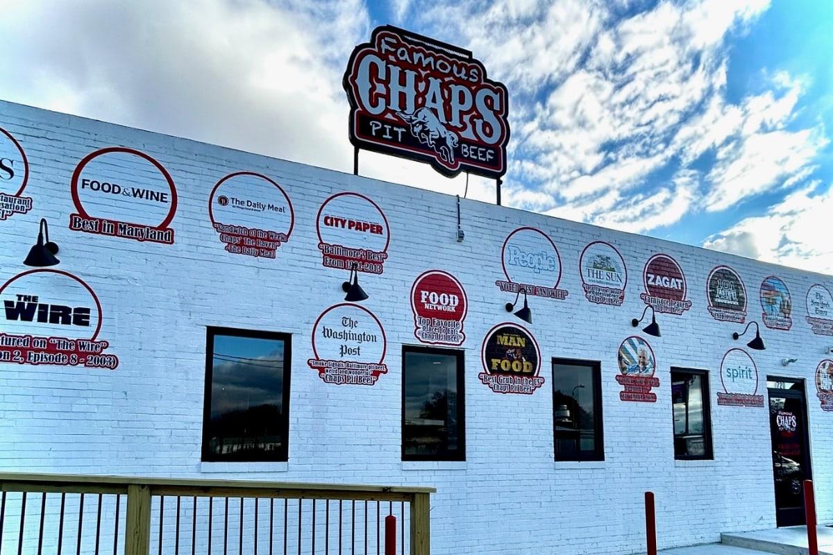 The white brick wall of Chaps Pit Beef under its neon sign, painted with the seals of the press that has written it up, including one for The Wire
