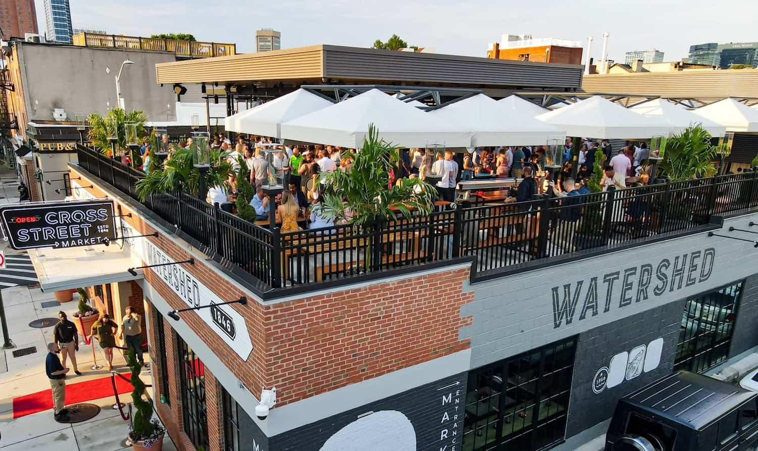 Watershed's roof deck above Cross Street Market, full under white umbrellas, with the market's neon sign on the corner below