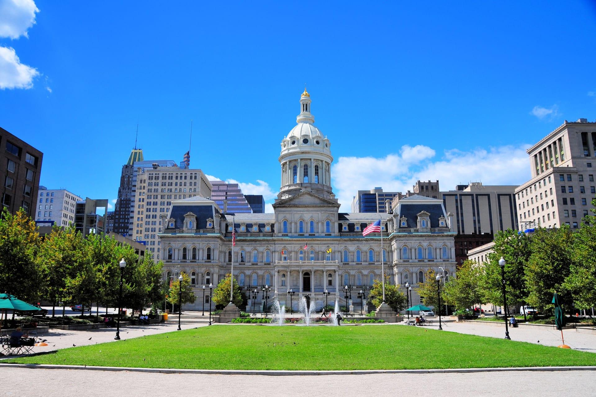 Baltimore City Hall and its dome across the lawn of War Memorial Plaza, office towers either side, under a blue sky
