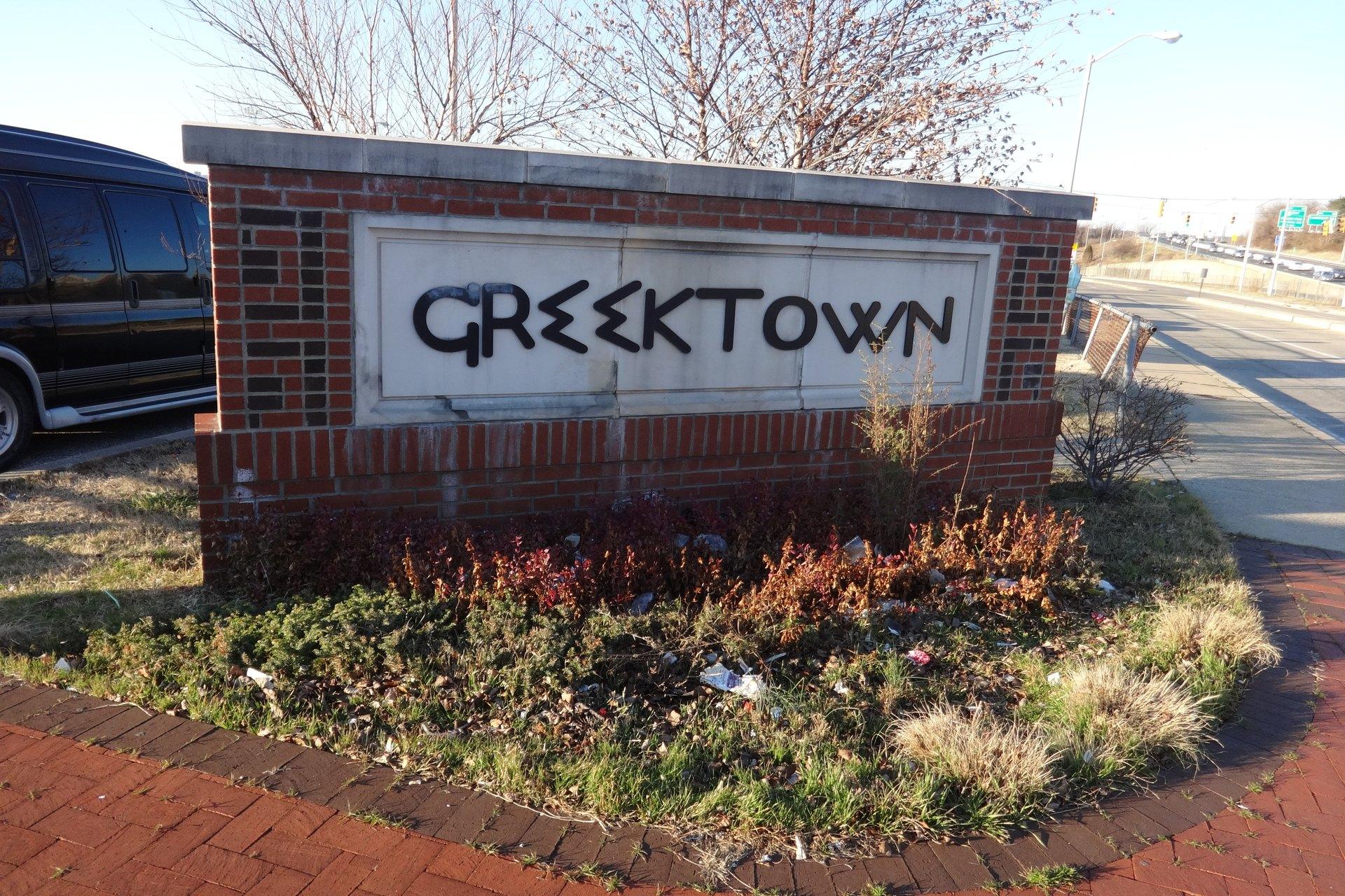 A brick and stone marker reading GREEKTOWN beside a road, bare trees behind it and the highway running away to the right