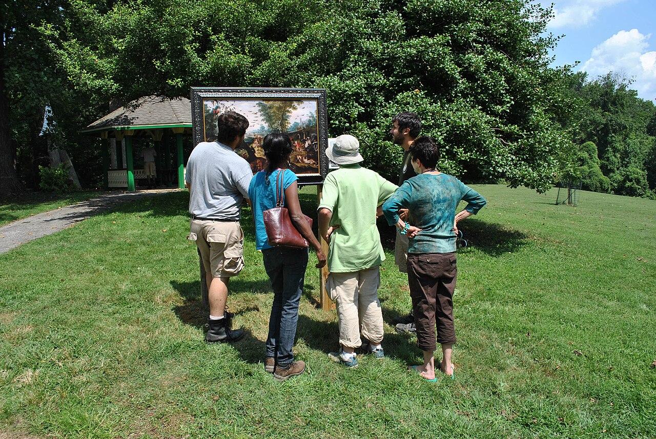 Visitors on the lawn in Gwynns Falls Leakin Park looking at a framed painting set up on an easel, trees and a pavilion behind
