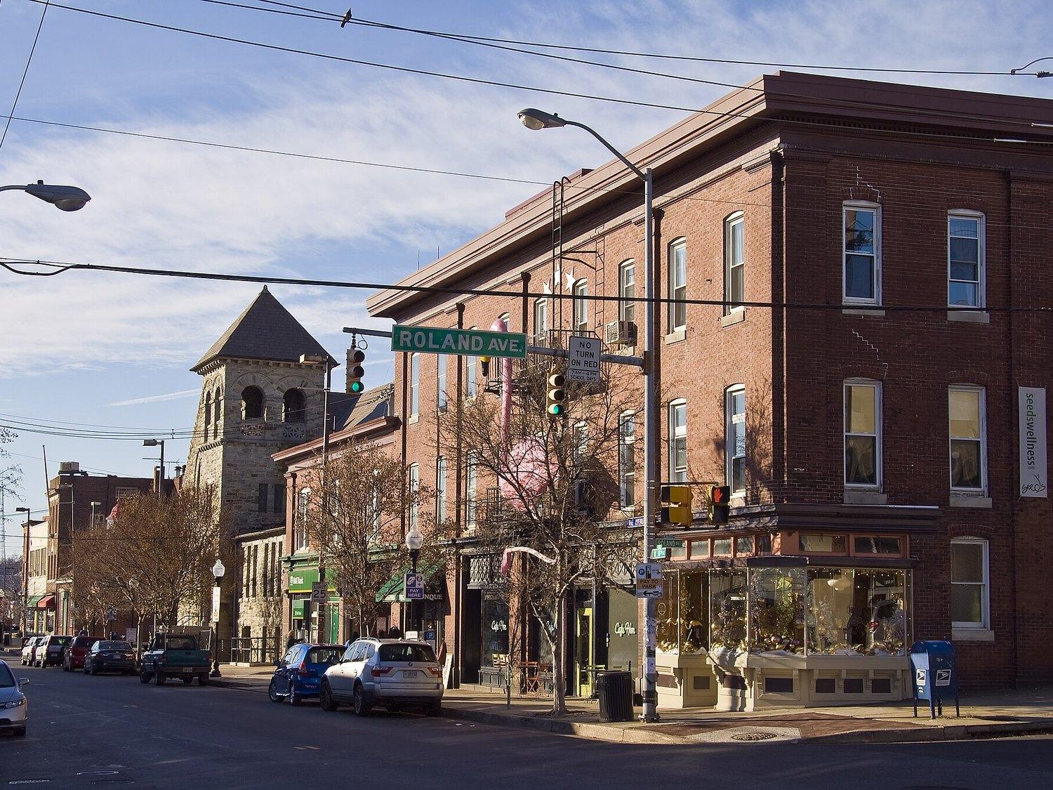 The Avenue at 36th and Roland, shopfronts along the brick corner block with the stone church tower behind