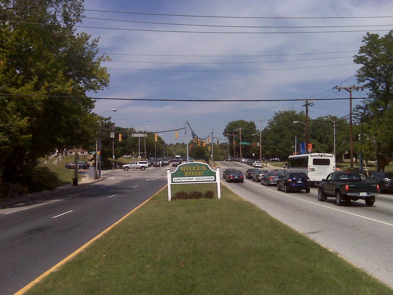 Hillen Road from its grassy median in front of Morgan State University, a Hillen Road Improvement Association sign, traffic lights at Cold Spring Lane ahead and a Morgan State shuttle bus on the right, photographed in 2010