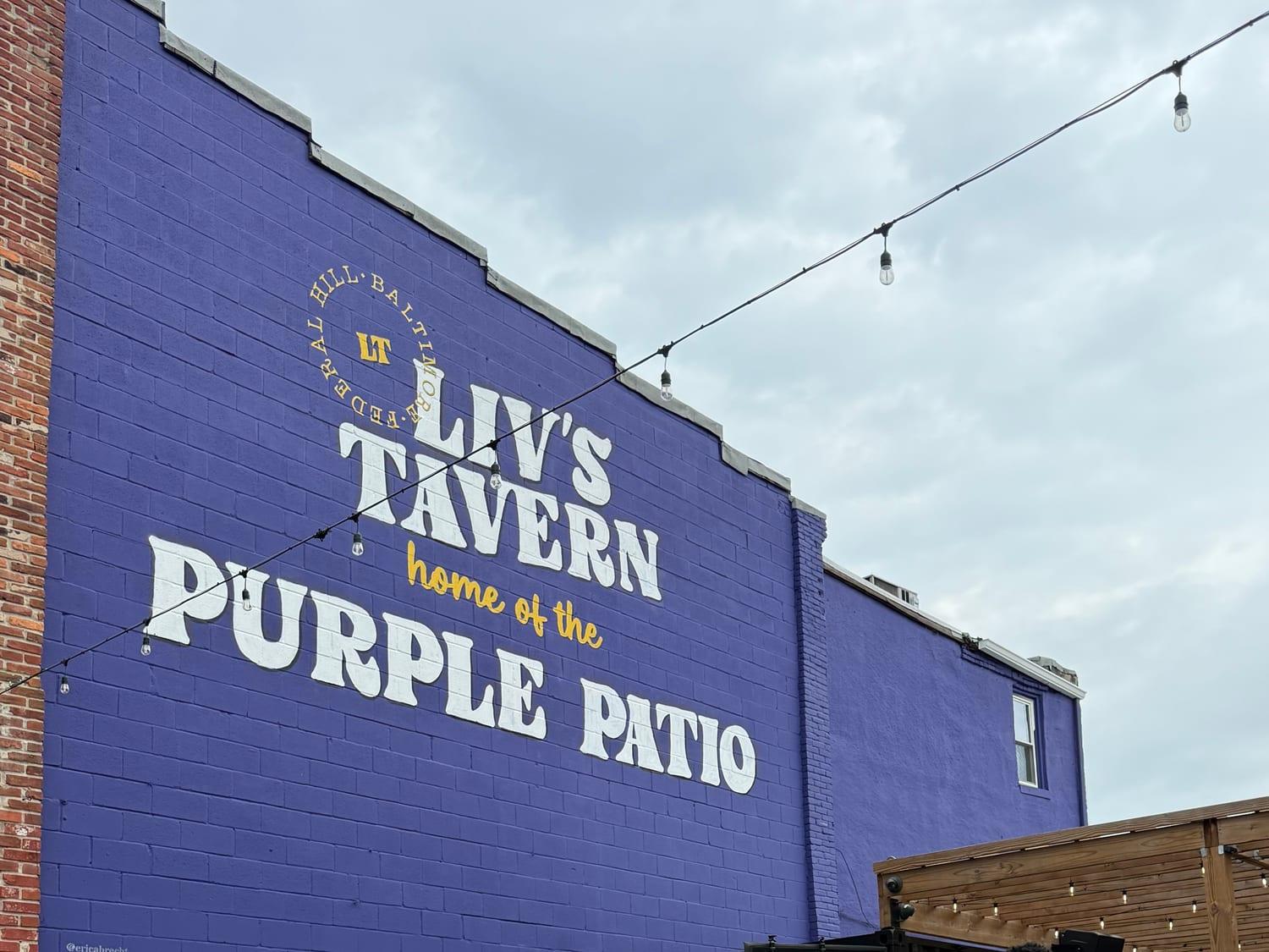 The painted wall at Liv's Tavern reading LIV'S TAVERN, home of the PURPLE PATIO, with string lights across the yard