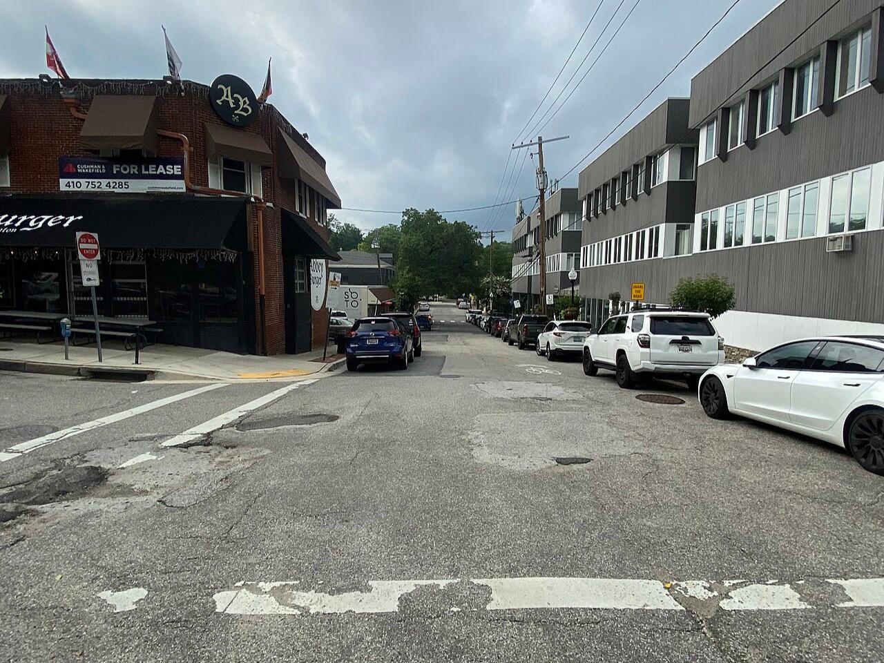 A street in Mount Washington village, low brick and clapboard shopfronts, parked cars and trees beyond