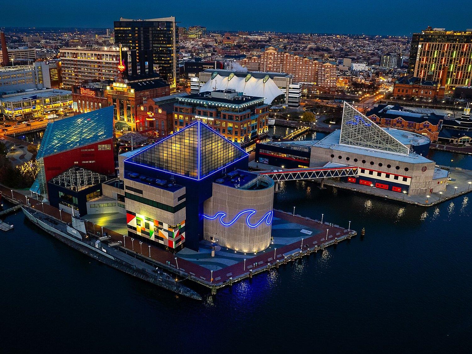 The National Aquarium lit blue at night on its Inner Harbor pier, seen from the air with the city behind