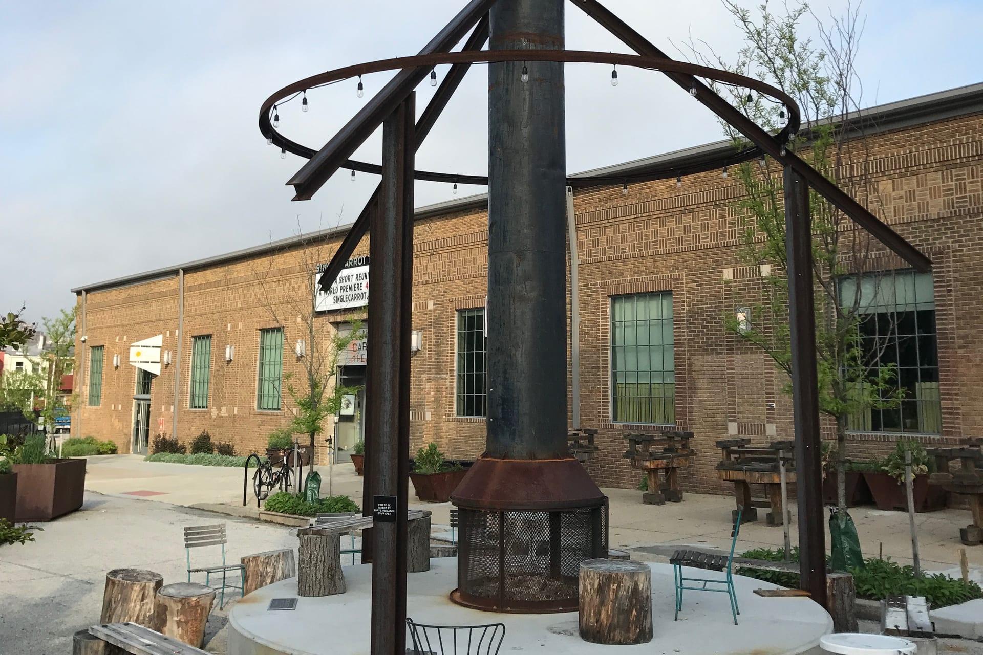 The Remington Community Plaza, its steel chimney strung with lights over café chairs and log stools, the brick mill building behind