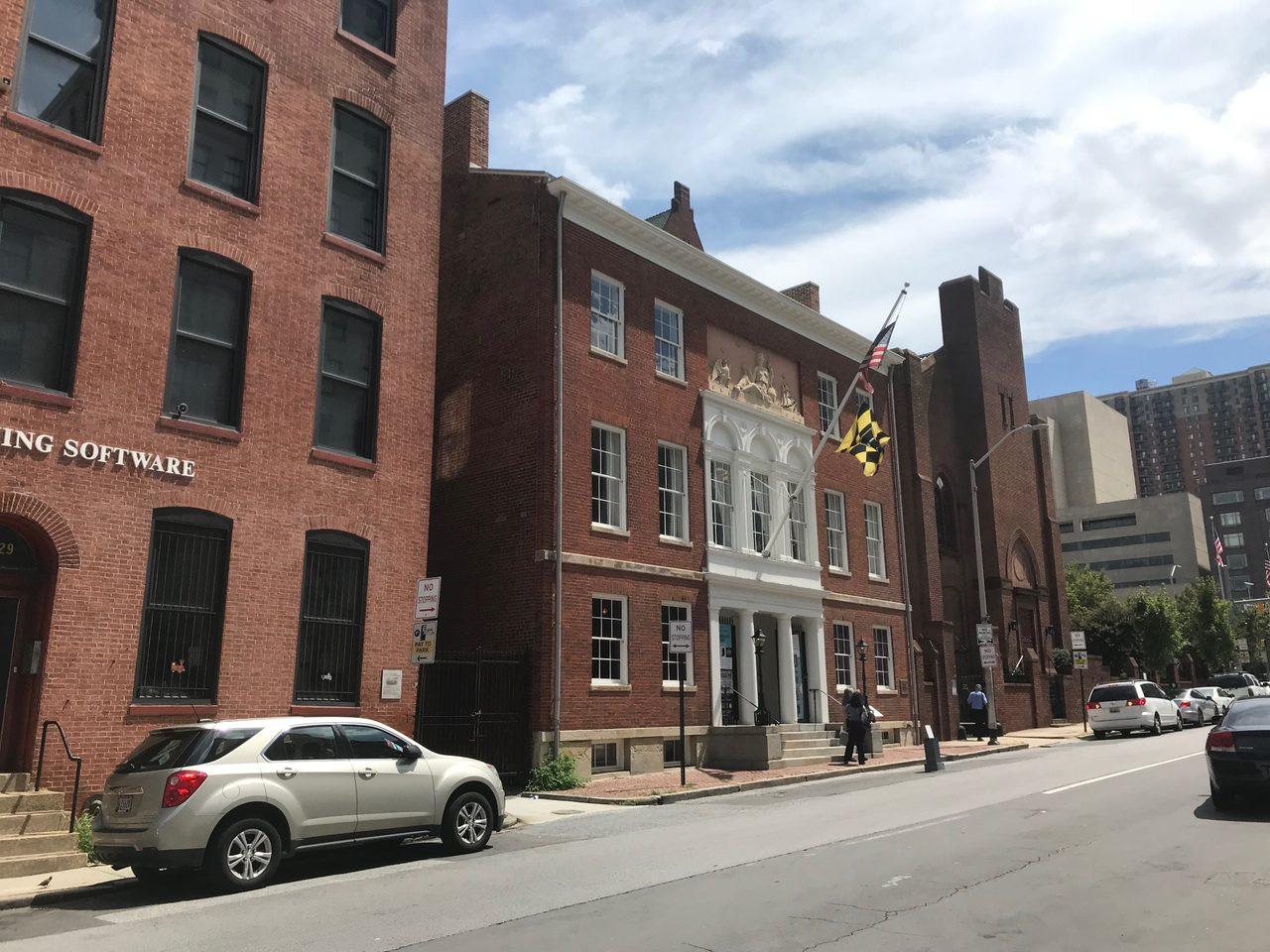 The Peale on Holliday Street, a three storey red brick museum building with a white arched portico, a carved relief above it and the Maryland flag flying
