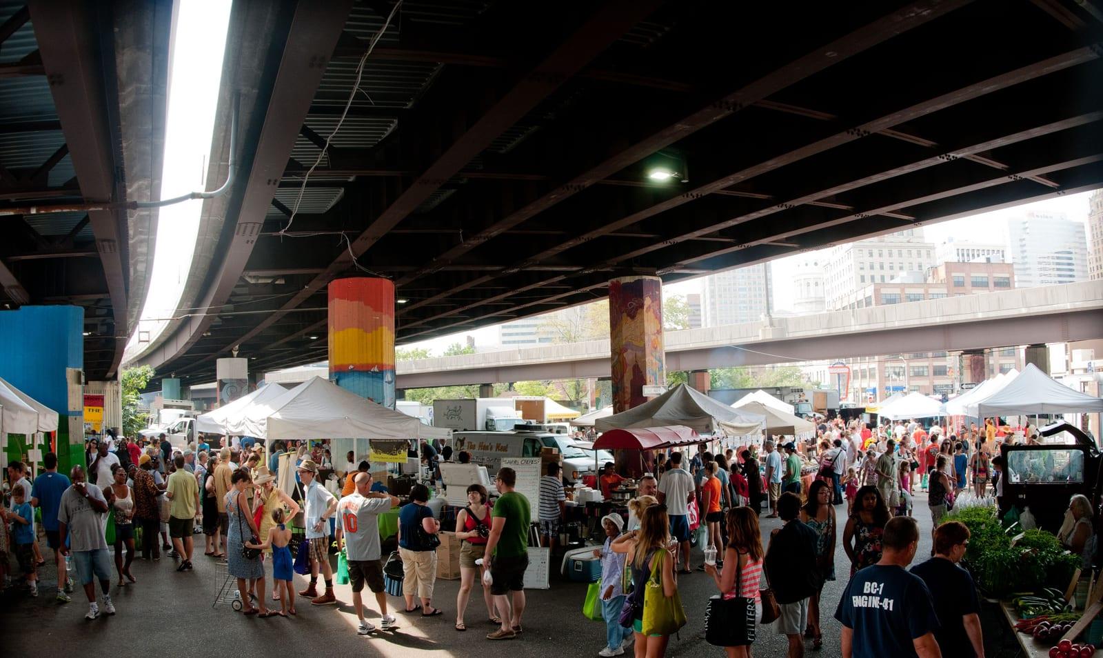 White market tents and a Sunday crowd in the car park under the Jones Falls Expressway, painted pillars holding up the road and downtown towers beyond, photographed in 2012