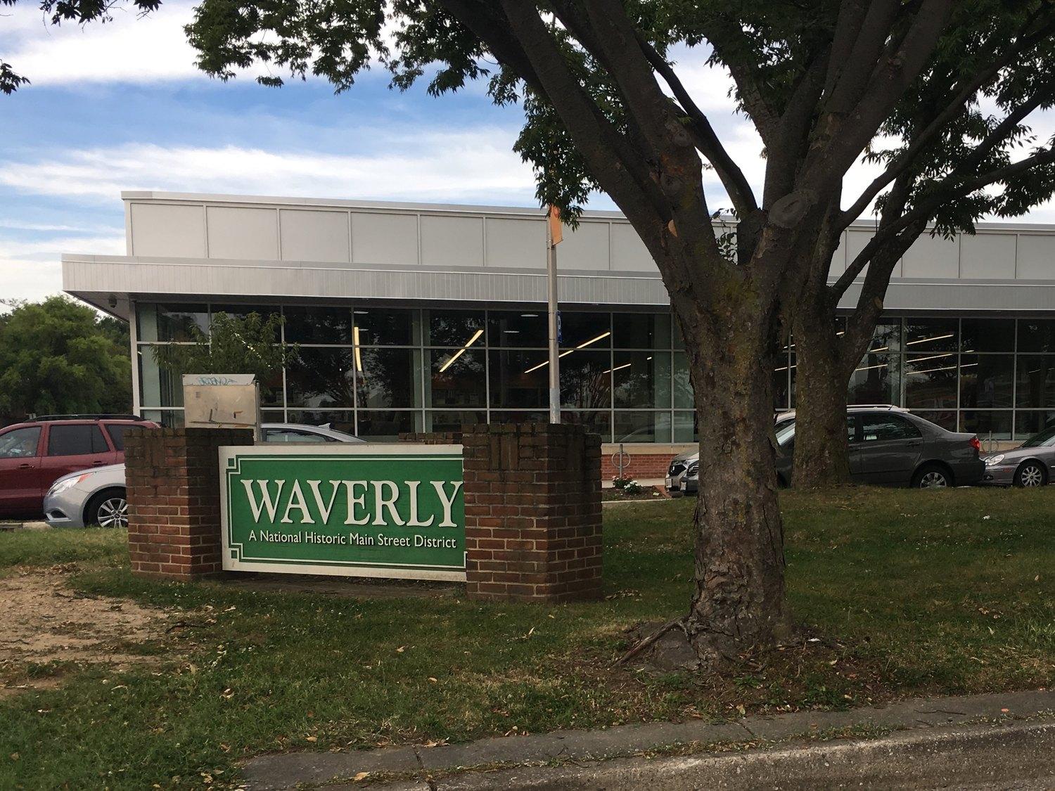 The green Waverly sign on its brick plinth, naming the National Historic Main Street District, with the shopfronts of Greenmount Avenue behind